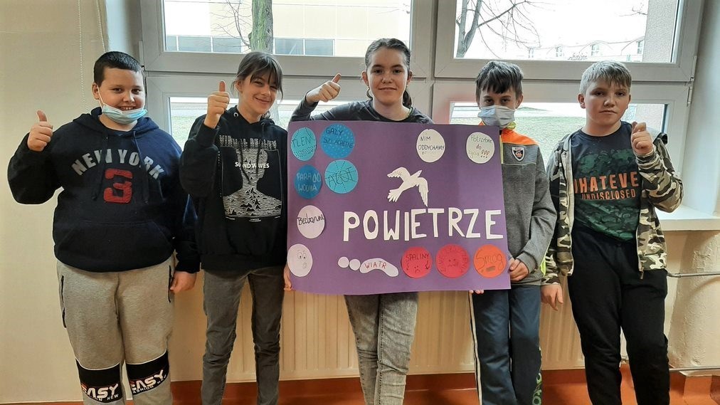 Grupa uczniów stojących pod oknem trzymający w dłoniach plakat.