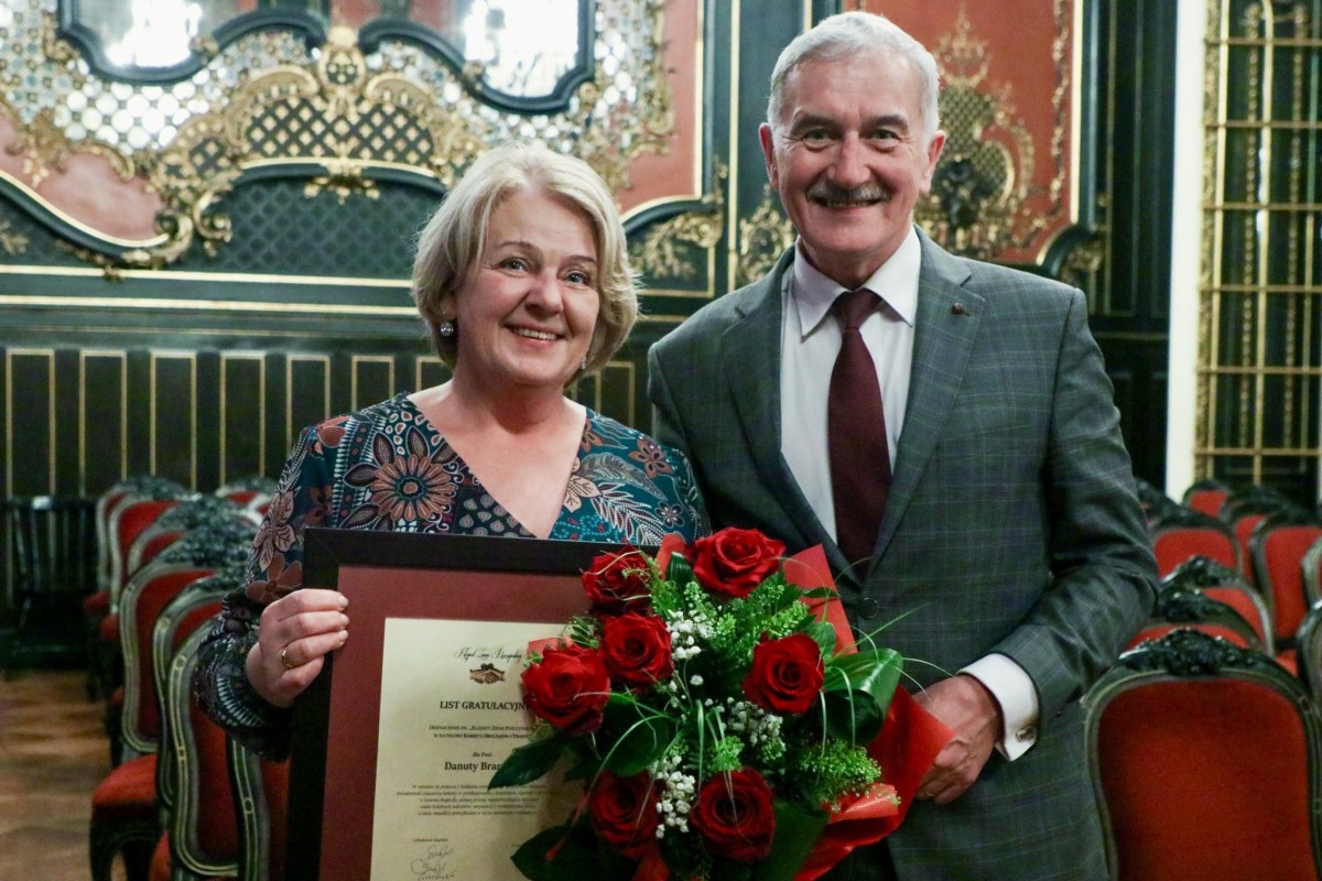 Kobieta stoi obok członka Zarządu Grzegorza Nogłego, trzyma w ręku kwiaty i tablice po uroczystej gali klejnot ziemi pszczyńskiej w sali lustrzanej.