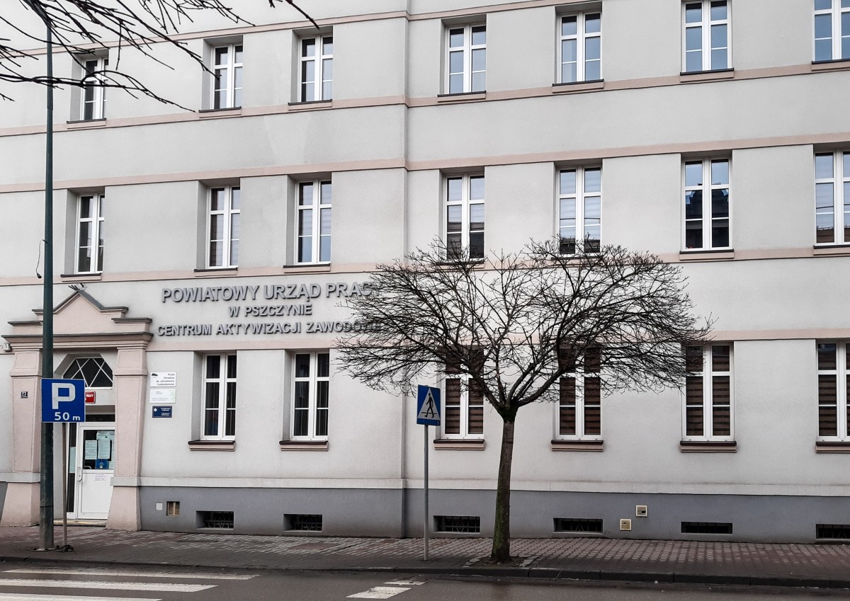 Zdjęcie przedstawia drogę, przy której stoi wysoki budynek z dużą ilością okien. To budynek Powiatowego Urzędu Pracy w Pszczynie. Przed nim drzewo bez liści.