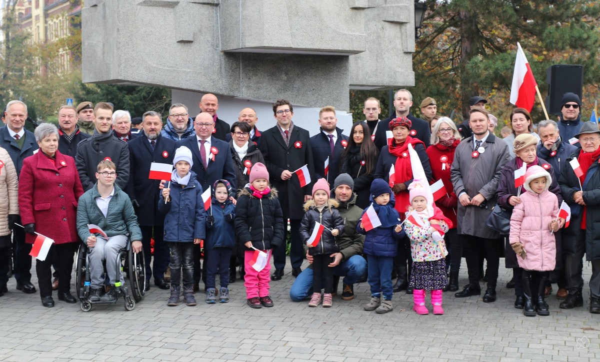 Grupa uczetsników uroczystości stoi z flagami przed pomnikiem