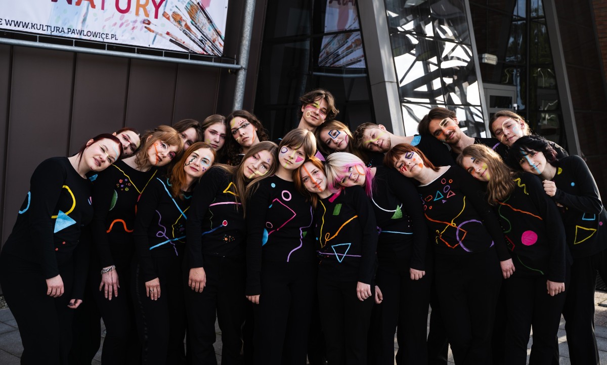 Członkowie grupy musicalowej ubrani w czarne stroje z kolorowymi wstawkami, pozują do zdjęcia, przytulając się do siebie
