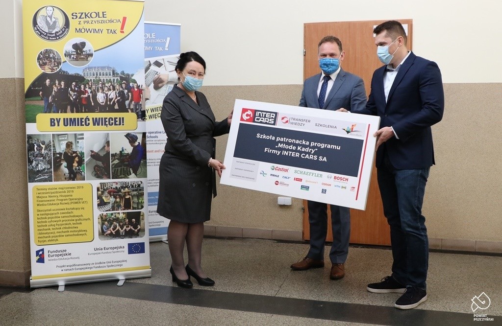 Dyrektor szkoły, wicestarosta i przedstawiciel firmy Inter Cars przekazujący tablicę szkoły patronackiej w budynku warsztatów szkolnych.