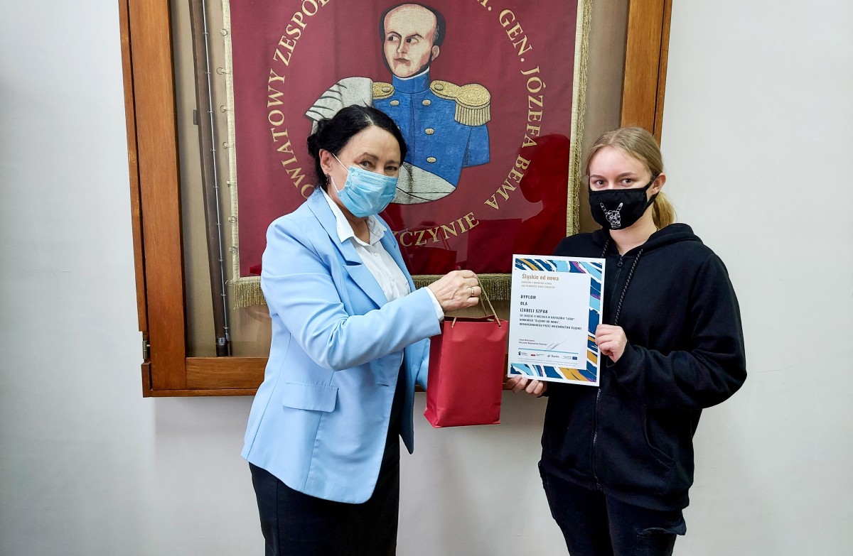 Dziewczyna w maseczce na twarzy trzyma dyplom, obok dyrektor PZS nr 1 w Pszczynie. Za nimi gablota ze sztandarem szkoły.