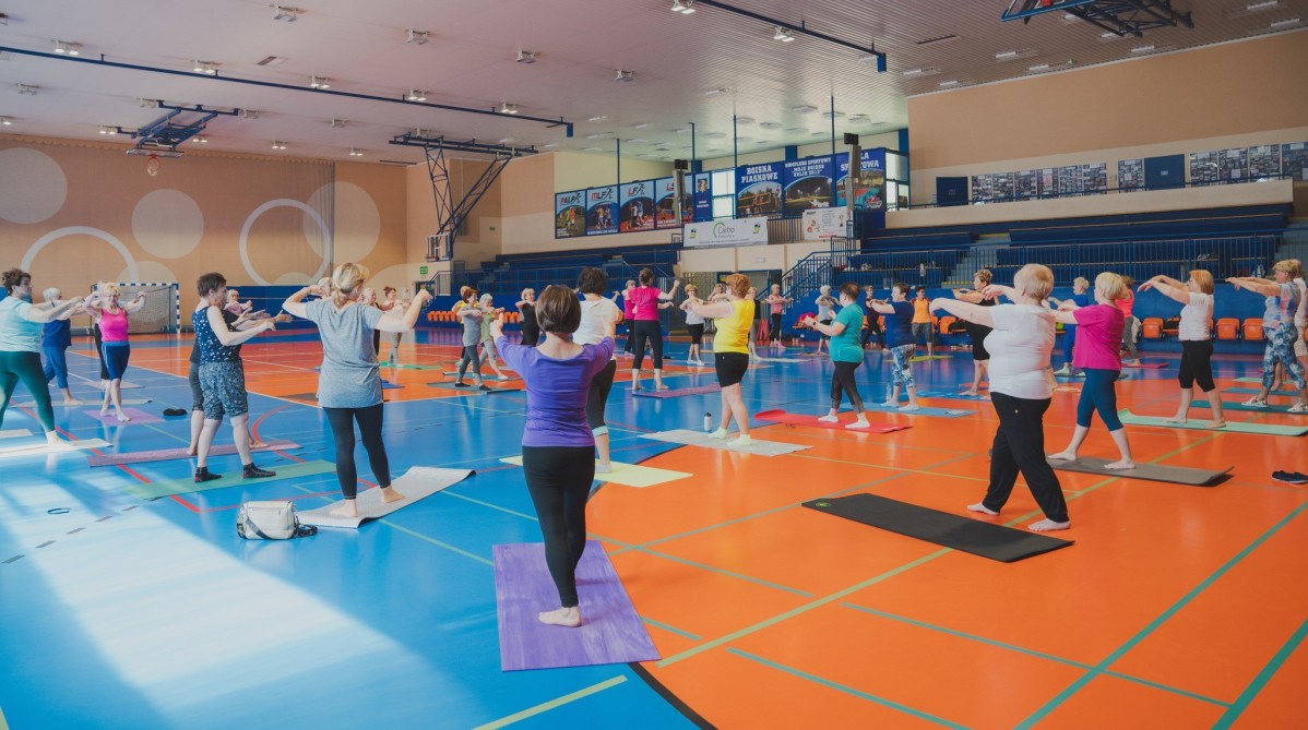 W hali ośrodka sportu trwają zajęcia gimnastyczne dla seniorów. Seniorzy ubrani w stroje sportowe wykonują ćwiczenia, stojąc na matach