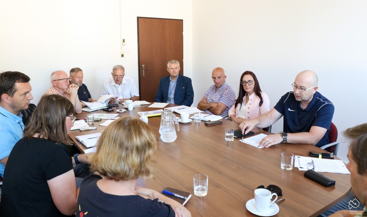 Grupa osób siedzi przy owalnym stole, na którym położone są dokumenty, rozmawiają.