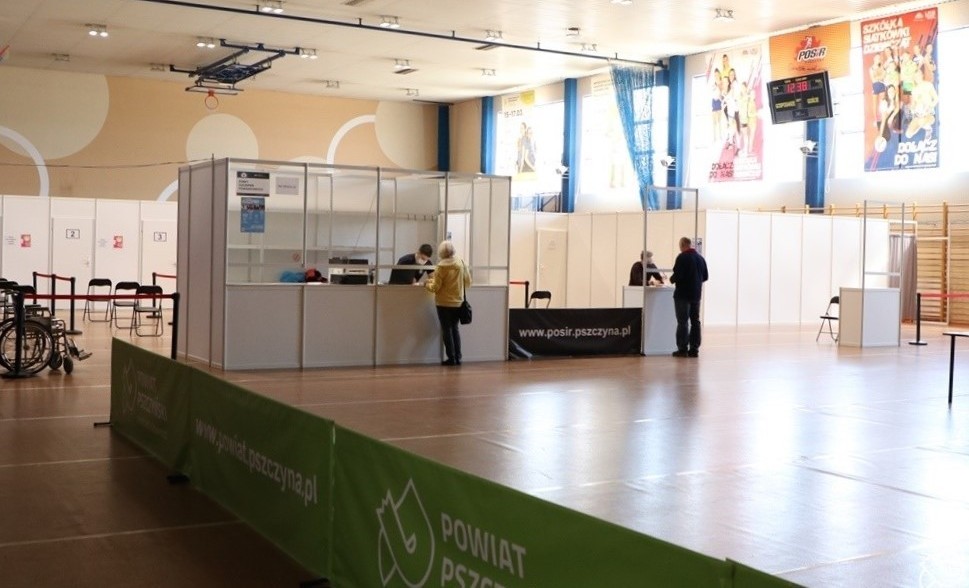 Wnętrze hali sportowej w Pszczynie, przeorganizowanej na punkt szczepień powszechnych. Na pierwszym planie zielone banery z logiem powiatu, na środku punkt informacyjny i stanowiska do szczepień.