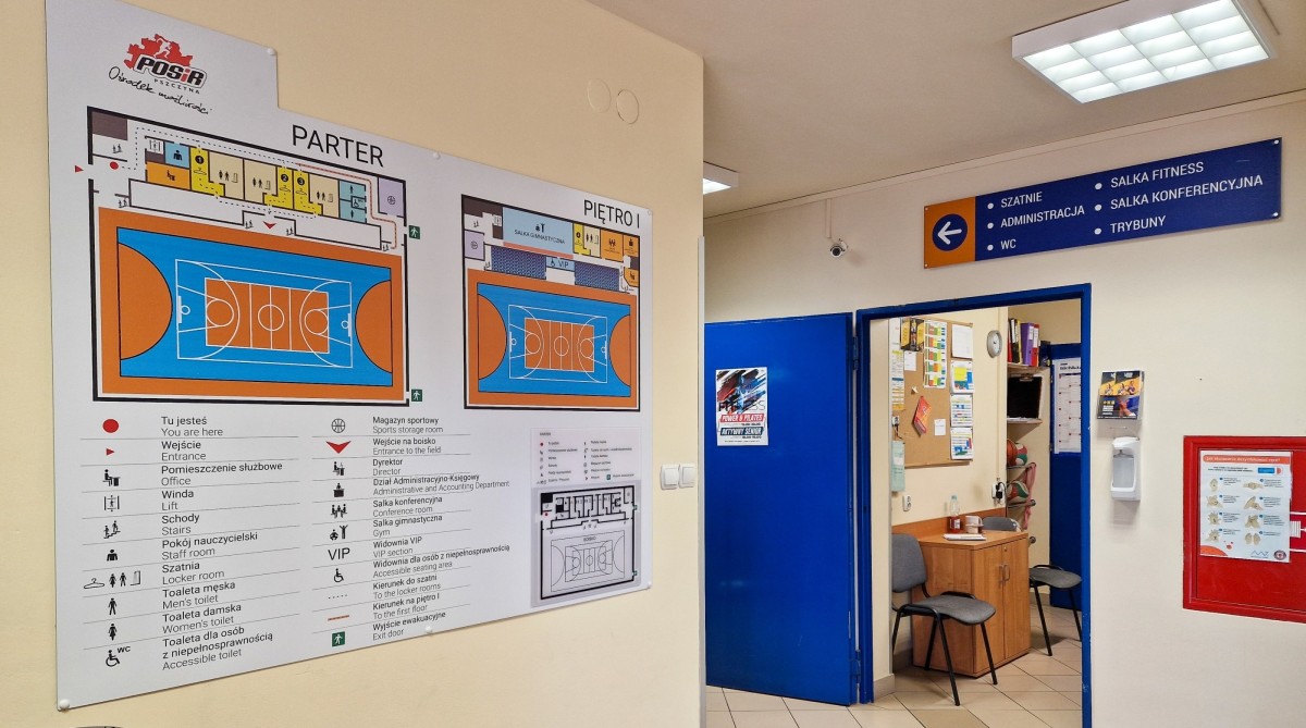 korytarz w hali Powiatowego Ośrodka Sportu i Rekracji. Na ścianie wisi duża tablica informacyjna z udogodnieniami dla osób ze szczególnymi potrzebami.