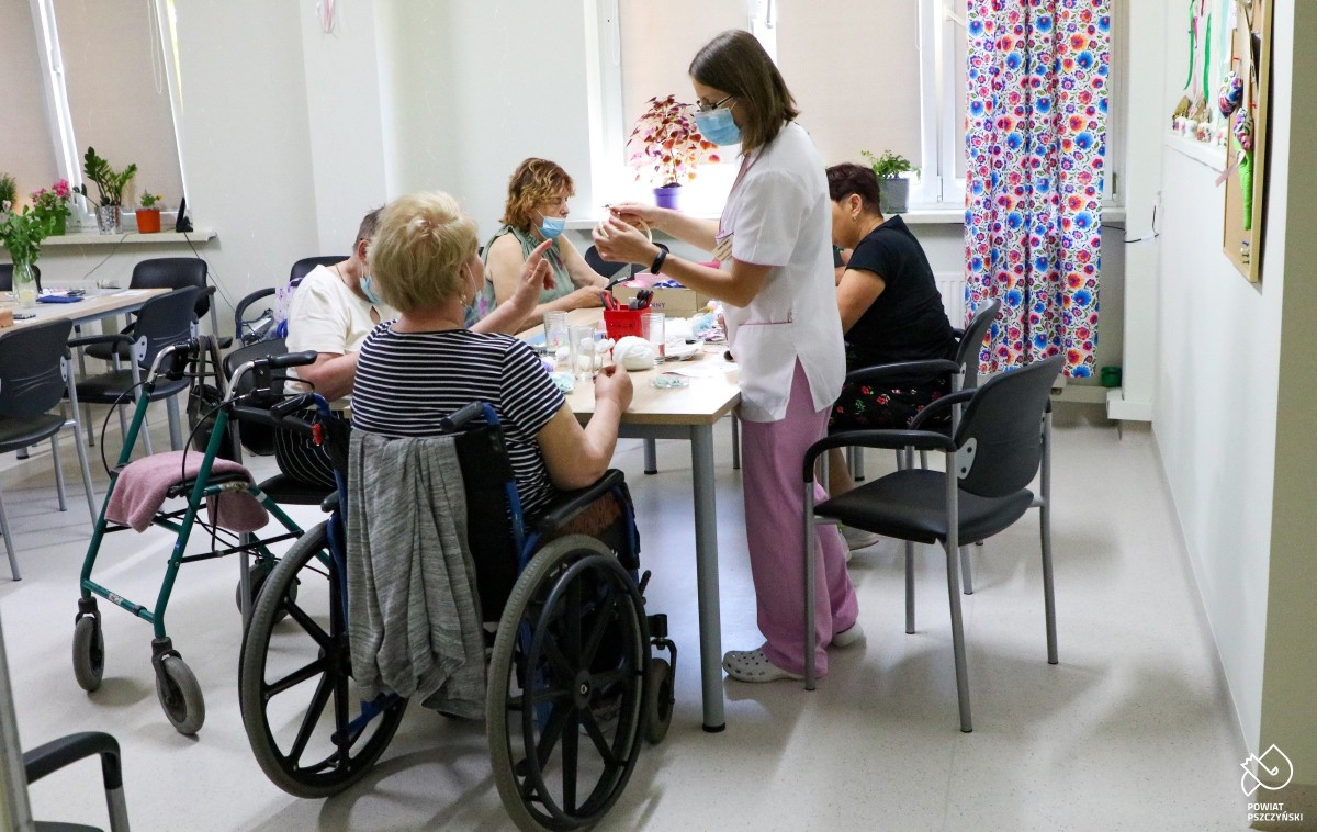 Pielęgniarki wraz z pacjentami DDOMU wewnątrz jasnej sali. Siedzą przy stolikach, wykonują prace.
