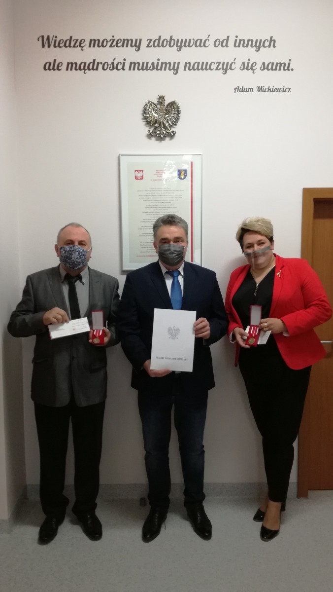 Grafika przedstawia troje nauczycieli którzy stoją w pomieszczeniu, od lewej dóch mężczyzsn i kobieta, w rekach trzymają medale i nagrody otrzymane z okazji Dnia Edukacji Narodowej.