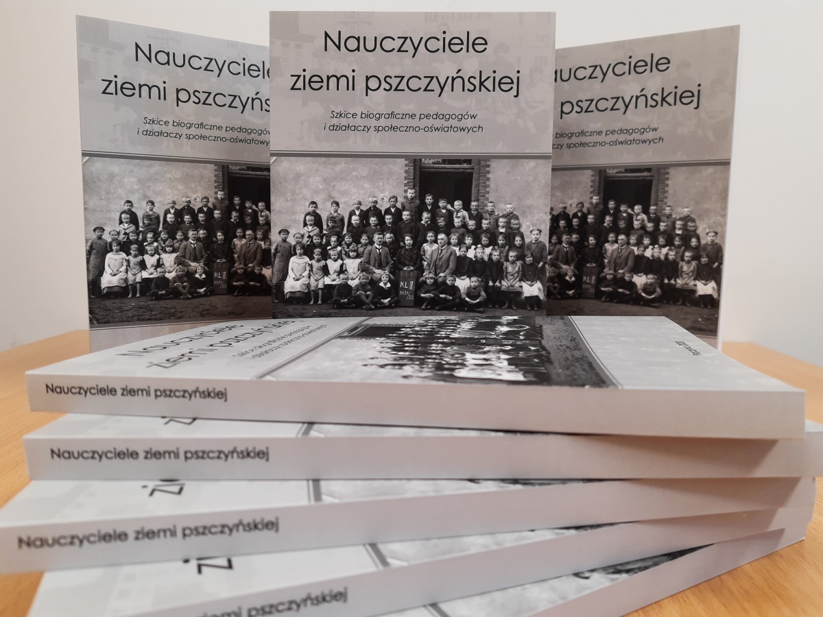 Na stole położone są książki. Za nimi, trzy książki ustawione są w pionie. Na okładce książki stara fotografia uczniów.