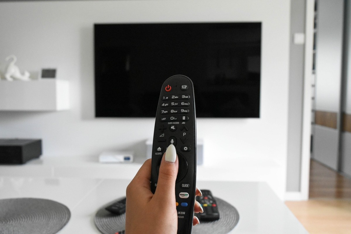 Dłoń trzyma pilot do telewizora. Na drugim planie telewizor