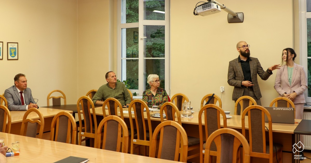 Kilka osób siedzących za stołem, liderka zespołu stoi i prezentuje wyniki ankiet, mała sala pszczyńskiego starostwa.